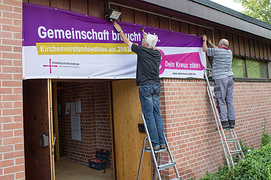 Heinz Rau und Erwin Henkel stehen auf Leitern vor dem Eingang der Trinitatiskirche und bringen ein Transparent zur Kirchenvorstandswahl an.