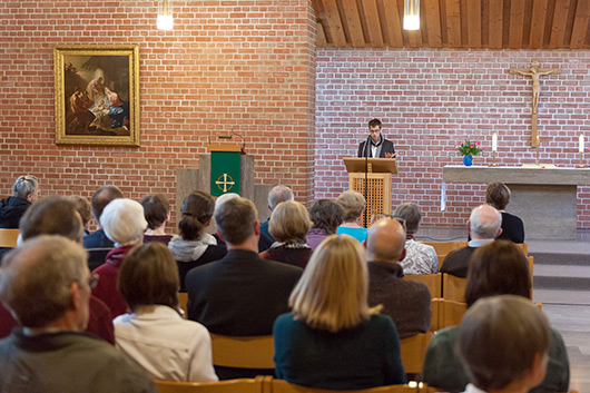 Blick aus dem hinteren Teil der Kirche in Richtung Altarraum; am Lesepult Sebastian Fix, im Vordergrund die Besucherinnen und Besucher des Vortrags.