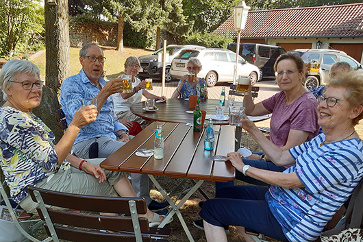 Teilnehmer*innen des Gemeindeausflugs sitzen im Freien an einem Tisch und prosten dem Fotografen zu.