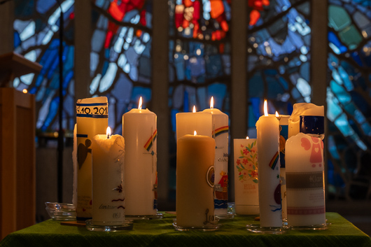 Entzndete Kerzen stehen auf einem flachen Tisch im Altarbereich der Trinitatiskirche. Im Hintergrund ist unschaft das Heiliggeist-Fenster der Trinitatiskirche zu sehen.